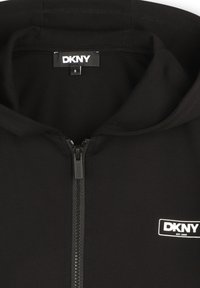 Haut zippé noir fabriqué en tissu doux, avec une étiquette logo à l'intérieur du col et un zip sécurisé. Design simple et épuré.