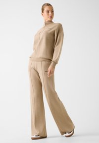 Beige trainingsset bestaande uit een sweatshirt met hoge kraag en broek met wijd uitlopende pijpen. Gladde stof met minimalistische branding aan de zijkant.