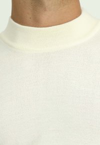 Weißer gerippter Strickpullover mit hohem Kragen und weich-texturierter Oberfläche, bei dem die engen Nähte im gesamten Stoff sichtbar sind.