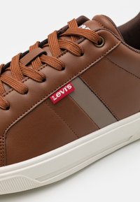 Bruna syntetiska läder sneakers med bruna snörningar, har en röd Levi's etikett och en texturerad vit gummisula, accentuerad med ett grått streck.