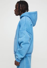 Lyseblå fleecehette med romslig hette, frontlomme og ribbestrikkede mansjetter. Myk tekstur, avslappet passform og litt oversized design.