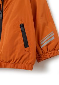 Chaqueta naranja con cierre frontal, puños elásticos y dos bolsillos negros con cremallera. Las franjas reflectantes en las mangas mejoran la visibilidad.