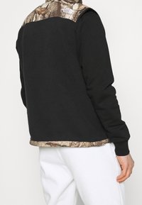 Czarny sweter z polaru z kamuflażowym wzorem na górnej części pleców; ma okrągły dekolt i teksturowany materiał; ściągacze w mankietach i na dolnym brzegu.