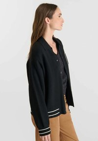 Cardigan nero con collo a camicia, dotato di tre bottoni, strisce bianche ai polsini e all'orlo, e una vestibilità rilassata. Texture liscia.