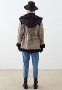 Persona con abrigo de cuadros con ribete de piel sintética negra, sombrero negro, guantes negros, vaqueros azules y botas negras hasta el tobillo de espaldas sobre un fondo claro.