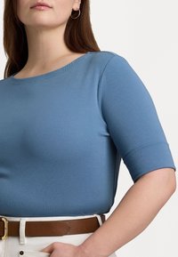 Ljusblå, figurnära topp med korta ärmar, gjord av ett slätt material. Har en rundad halsringning och en subtil textur. Kombinerad med ett brunt bälte.