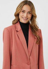 Blazer rosa estructurado con solapas de muesca, cierre de un solo botón y dos bolsillos frontales. Llevado sobre una parte superior de cuello alto negra. Textura de tela suave.