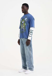 Blaues Grafik-T-Shirt mit Totenköpfen und floralem Design, kombiniert mit einem weißen Langarmshirt. Abgewetzte hellblaue Jeans mit Fransen.