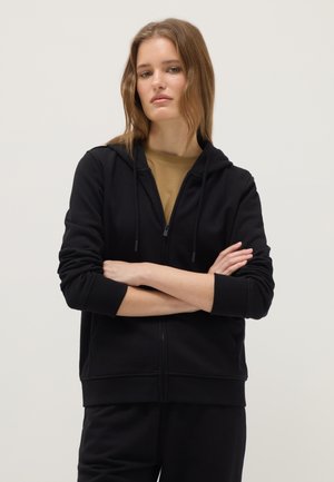 Junge Frau mit hellbraunen Haaren, die einen schwarzen Kapuzenpullover mit Reißverschluss trägt und die Arme vor einfachem Hintergrund verschränkt hat.