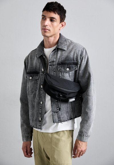 Veste en denim grise au design cropped, t-shirt blanc en dessous et pantalon beige. Un sac bandoulière noir avec des détails en mesh pend de manière décontractée.
