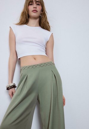 Junge Frau trägt ein weißes ärmelloses Crop-Top, olivgrüne Hosen mit weitem Bein und Kettengürtel sowie klobige Armbänder an einem Handgelenk.