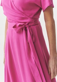 Pink wrap dress featuring short sleeves, a tied waist, and a flowing A-line skirt with a smooth texture and subtle stitching detail.