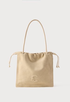 Τσάντα ώμου από μπεζ σουέντ με πλευρικά κορδόνια, μία μακριά λαβή και ένα διακριτικό ανάγλυφο λογότυπο στο κέντρο μπροστά.