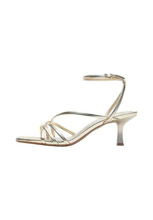 Guld sandaler med smalle stropper, lav killingehæl, ankelrem med spænde, åben tå og minimalistisk design mod hvid baggrund.