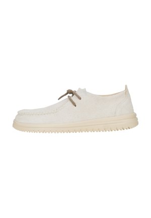Scarpe in suede color crema con punta tondeggiante, caratterizzate da un design con lacci e una lace marrone a contrasto, poggiate su una suola beige chiaro.