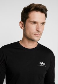 Schwarz langärmliges T-Shirt aus Baumwolle mit einem kleinen weißen Logo auf der linken Brustseite und einem runden Halsausschnitt. Glatte Textur.