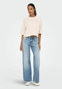 Frau in einem cremefarbenen, kurz geschnittenen Sweatshirt, hellblauen weit geschnittenen Jeans, weißen Sneakers und mit großen Ohrringen, die vor einem schlichten weißen Hintergrund steht.