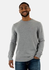 Maglione grigio lavorato a maglia con scollo rotondo, polsini e orlo a coste. Presenta una piccola etichetta con logo sul petto. Indossato con jeans blu.