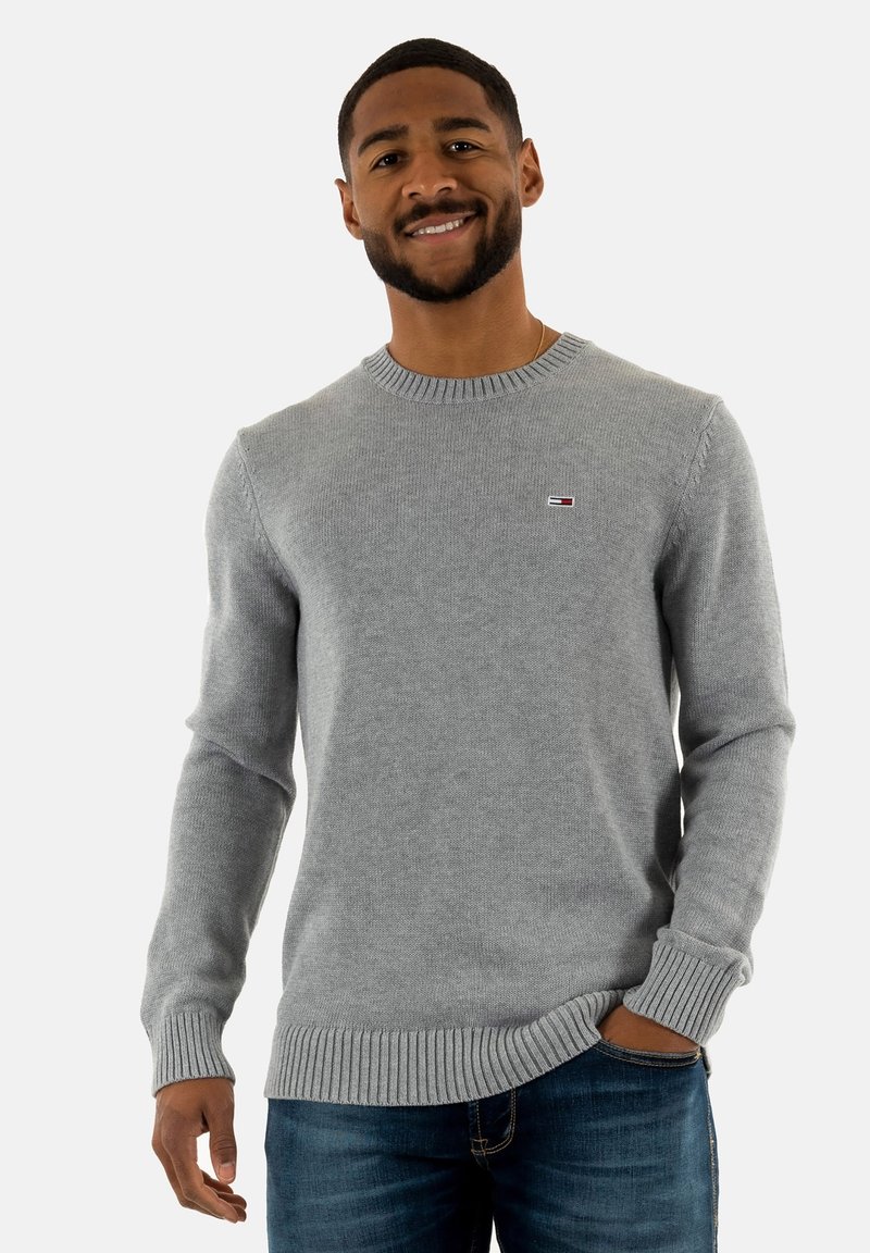 Maglione grigio lavorato a maglia con scollo rotondo, polsini e orlo a coste. Presenta una piccola etichetta con logo sul petto. Indossato con jeans blu.
