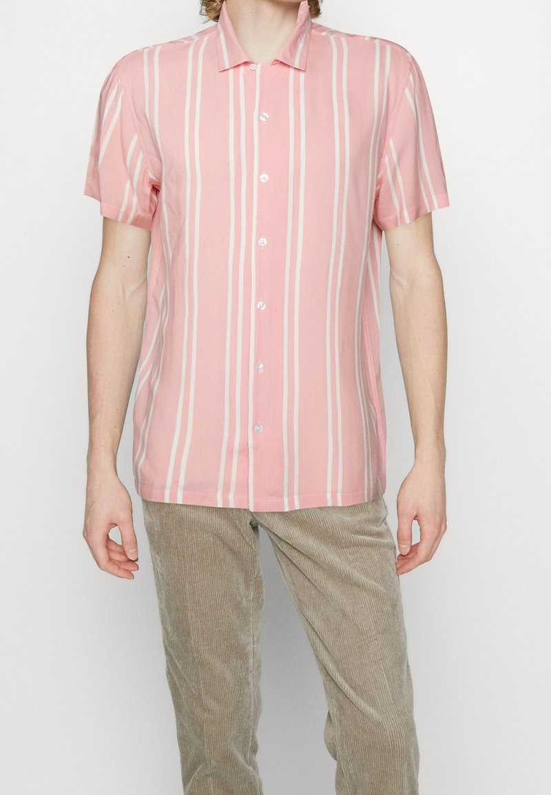 Homme portant une chemise rose à manches courtes à rayures blanches verticales et un pantalon beige en velours côtelé, debout sur fond blanc.