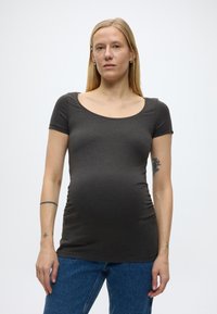 Terhes nő hosszú szőke hajjal, sötét színű, testhezálló rövid ujjú felsőt és kék farmert visel, egyszerű világos háttér előtt állva.