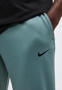 Κοντινό πλάνο ατόμου με τυρκουάζ παντελόνια Nike με μαύρο λογότυπο Swoosh, το χέρι ακουμπά στην τσέπη και μαύρο πουκάμισο ορατό στη μέση.