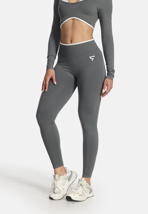 Žena nosí priliehavý sivý crop top s dlhým rukávom a zodpovedajúce vysoké legíny s bielymi teniskami na bielom pozadí.
