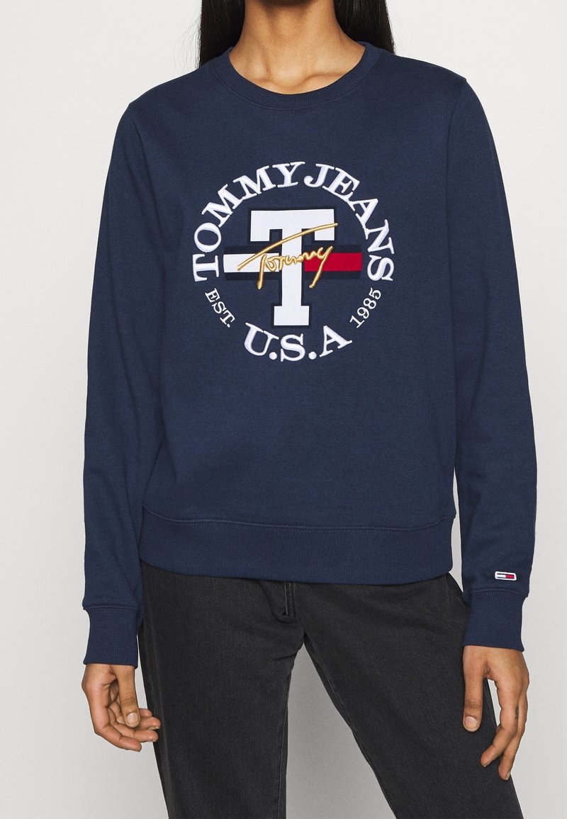 Sweat-shirt bleu marine avec un grand "T" blanc, le texte "Tommy Jeans U.S.A. Est. 1985", un drapeau rouge et blanc, et le script doré "Tommy" à l'avant.