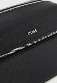 Juodas tekstūruotas odinis krepšelis su sidabro spalvos užtrauktuku ir įspaustu "BOSS" prekės ženklo logotipu priekyje, pasižymintis lygiu paviršiumi.