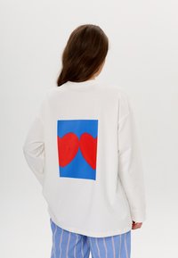 White oversized sweatshirt featuring a blue square design with two red hearts on the back. Soft fabric with long, loose sleeves.