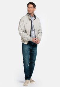 Lichtbeige bomberjack met een rits aan de voorkant, zwart accent, en een lichtblauw gebloemd shirt eronder, gecombineerd met donkerblauwe jeans en beige sneakers.