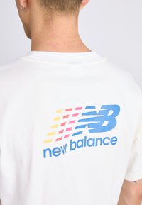 Tricou alb din bumbac cu un logo în gradient albastru și roșu, cu textul "new balance" dedesubt. Gât rotund, mâneci scurte, croială lejeră.