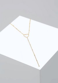 Collier délicat en or en forme de Y avec petits accents de perles et pendentif ouvert en forme de cœur, présenté sur une surface blanche inclinée.
