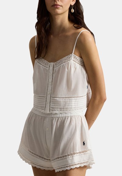 Woman wearing white sleeveless buttoned top and matching shorts with lace details and small embroidered logos on the side.