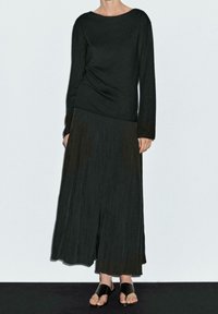 Musta pitkähihainen paita pyöreällä kaula-aukolla, yhdistettynä mustaan plissettyyn maksihameeseen ja mustiin litteisiin sandaaleihin. Tasainen, sileä pinta koko asussa.