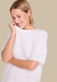 Hvit, myk strikkegenser med korte ermer og vid halsutskjæring. Lett, luftig tekstur med en liten fuzzy følelse og en avslappet passform.