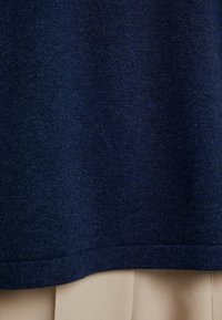 Maglione lavorato a maglia blu navy con tessuto texturizzato e orlo a coste. L'orlo si trova sopra un indumento di un leggero beige, mostrando colori contrastanti.