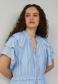 Hellblaues Kleid aus weichem Stoff mit Rüschensäumen, gerafftem Bund und V-Ausschnitt mit Bindeband. Glatte Textur und lässige Passform.