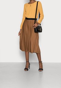 Top giallo a maniche lunghe con dettaglio a chiave, abbinato a una gonna plissettata marrone e tacchi a strappo neri, completato da una borsa nera.