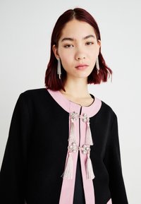 Cardigan tricotat negru, cu detalii roz și închideri cu toggle. Are ciucuri și mărgele decorative. Modelul poartă cercei lungi, strălucitori.