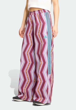 Personne portant un pantalon large avec des motifs zigzag violets et du texte jaune, des rayures bleu clair sur les côtés, associé à des baskets noires et un crop top blanc.