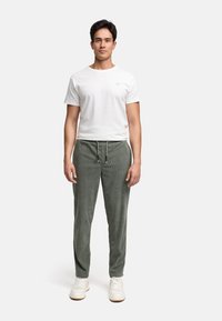 Mann, der nach vorne steht, trägt ein weißes kurzärmliges T-Shirt, olivgrüne Jogginghosen mit Kordelzug und weiße Sneakers vor einem einfarbigen Hintergrund.