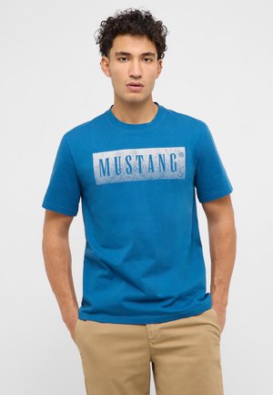 Mann mit lockigem Haar, trägt ein blaues Mustang-T-Shirt und beige Hosen, steht mit den Händen in den Taschen vor hellem Hintergrund.