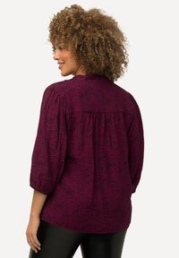 Bordeauxfarbene Bluse mit gepunktetem Muster, kurzen Puffärmelchen und einem gerafften Rückenpasse. Der Stoff wirkt leicht und strukturiert.