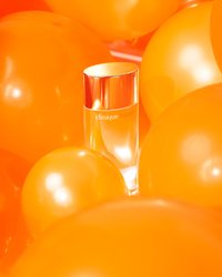 Duidelijke glazen fles van Clinique Happy parfum met een zilveren dop, omringd door heldere oranje glanzende ballonnen.