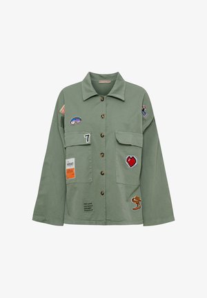 Olivengrøn jakke med knapper, brede ærmer, forlommer med klap og forskellige farverige patches, herunder et hjerte, tallet 7 og tegneseriefigurer.
