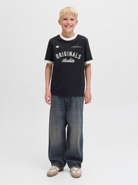 Tricou negru cu text alb „ORIGINALS studio”, guler de culoare deschisă, asortat cu blugi albaștri cu picioare largi și adidași bej.