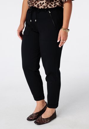 Vrouw die zwarte taps toelopende broek draagt met een zak met rits en een top met luipaardprint, gecombineerd met gouden armbanden en ringen en zwarte schoenen met een gaaspatroon.