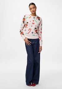 Kobieta w świątecznym swetrze z nadrukiem dziadka do orzechów, laski cukrowej, serduszek i płatków śniegu, w połączeniu z szerokimi niebieskimi dżinsami i czerwonymi butami.