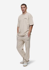 Beige bomulls t-skjorte med korte ermer og en svart logo på brystet, sammen med beige bukser. Begge plaggene har en avslappet passform.